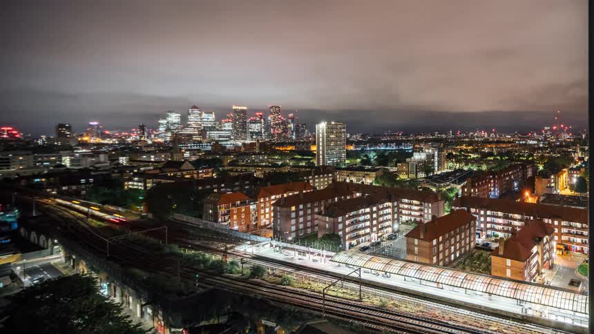 Trains pause while passing apartment buildings a distance from city skyline on a cloudy night