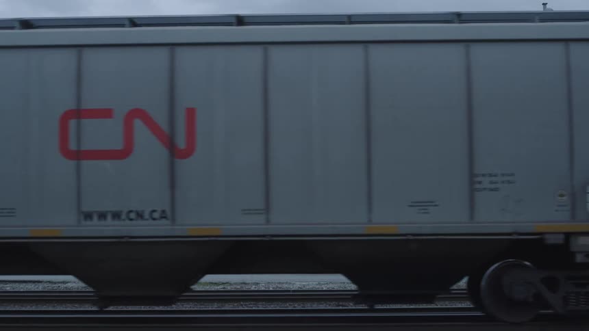 A train with containers and various written texts is moving on the tracks with trucks in the background