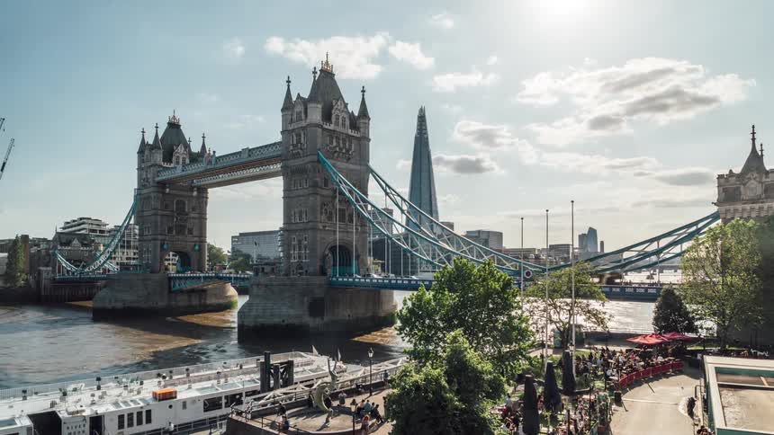 A busy london bridge with lots of cars going to and fro with a boat docked on shore as people walk by