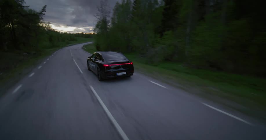 A black sports car is driving on an empty asphalt road and there are several trees on the side of the road
