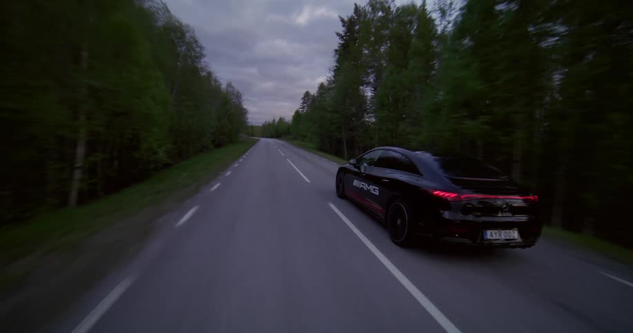 A black sports car is driving on an asphalt road and there are several trees outside the road