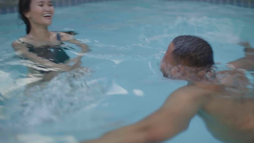 A man dives from the edge of a pool and comes to the surface next to a woman and they both smile