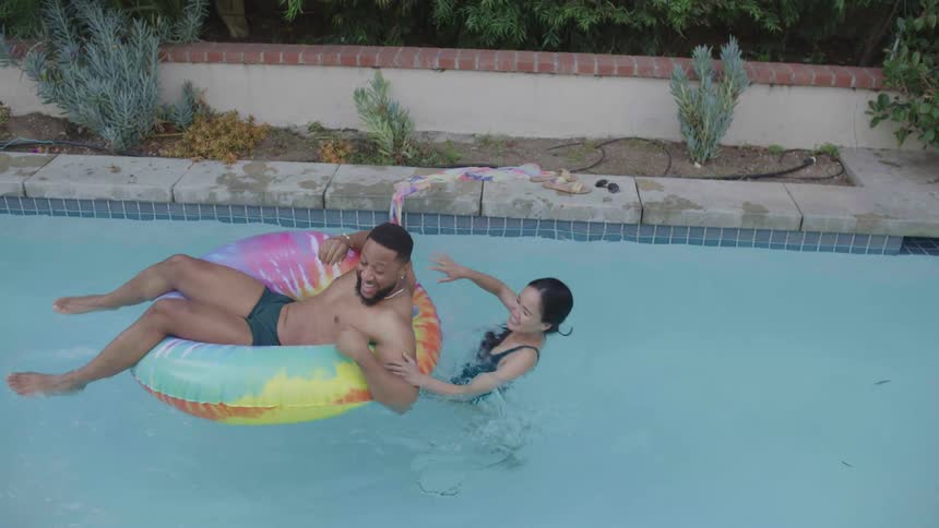 A man is on top of a buoy in a pool and a woman on the edge falls hugging the man and they are both happy