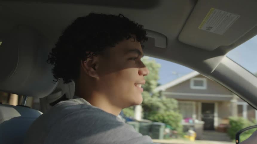 Man with curly hair and a gray t shirt drives a car listening to music