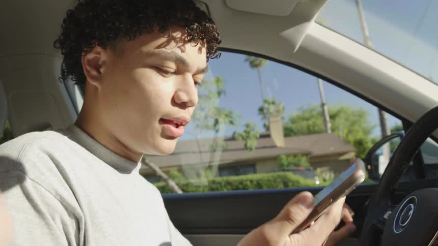A young man is holding a cell phone in his hand and talking while inside a car