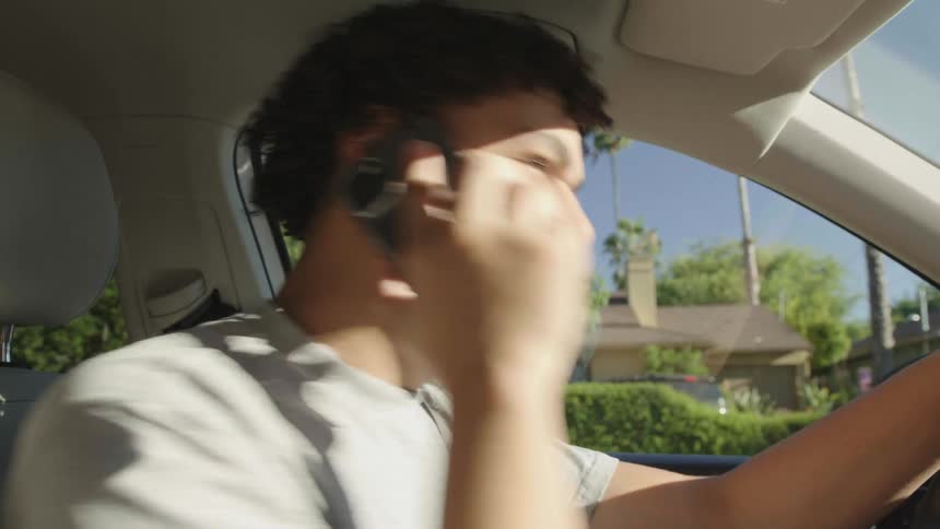 A young man driving a car pulls over to take a call looks behind him then takes off again after finishing the call