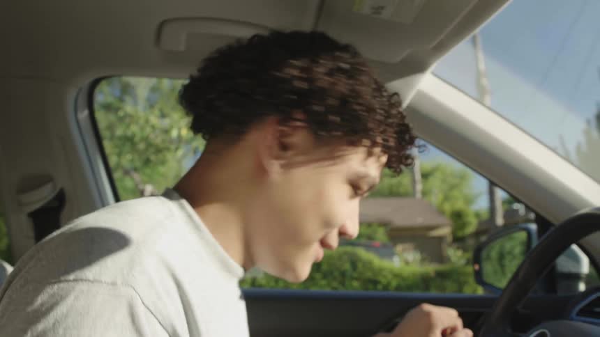 Teenager with curly hair a gray t shirt talks on speaker phone and gets out of car