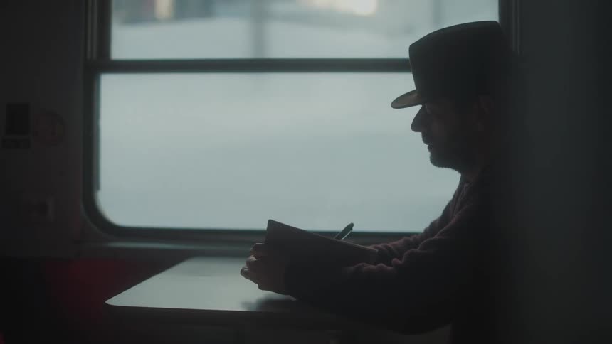 A man sits on a train wearing a bowler hat and writing in a notebook with a snowy ground through the train window