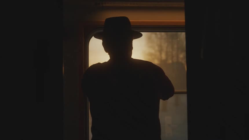 Silhouette of a man with his back turned and wearing a hat looking out of a train window