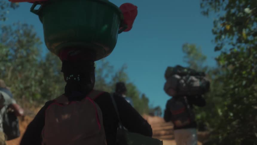 People walking with backpacks and a woman carrying a bucket on her head along a trail