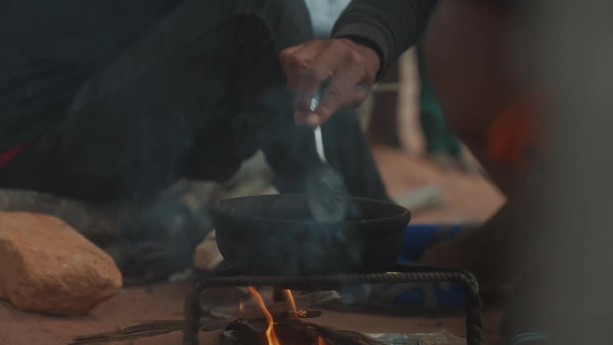 Closeup of a persons hand with a spoon preparing food in a pot over an open fire on the ground