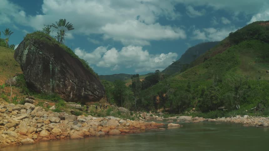 Hikers move near base of large boulder standing near shore and mountains