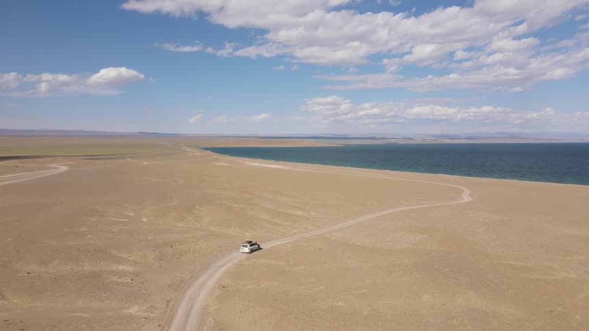 A small white car is driving on a dirt road in a desert and in front there is a sea