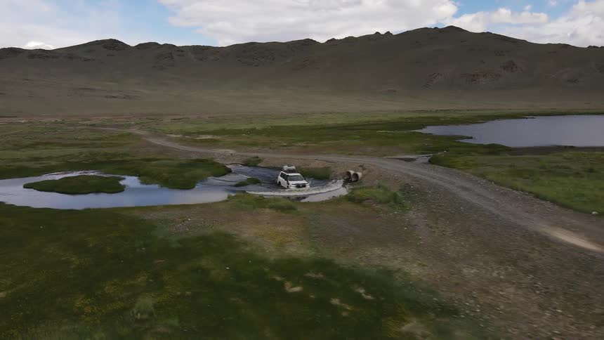 Vehicle parallels dirt road and drives through water with mountains in the background