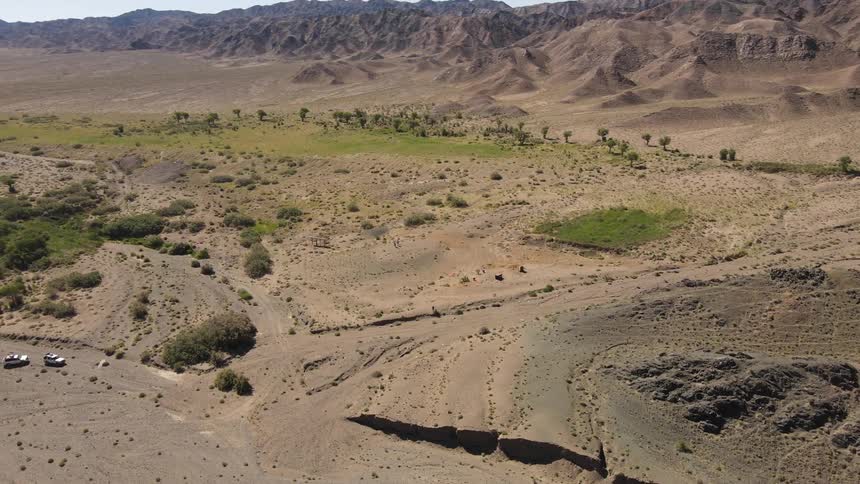 Low aerial view orbiting a hill with green vegetation on a hill in a desert valley with a jeep parked on a circular path and dunes in the background on a bright day