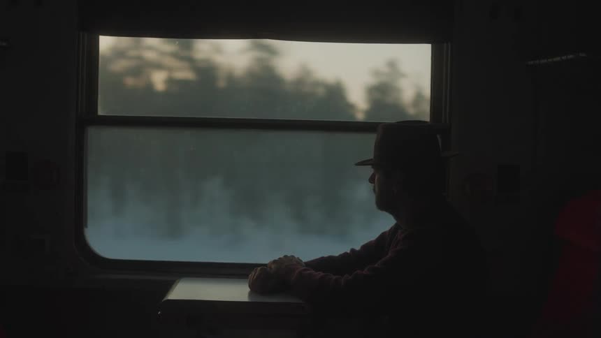 A man wearing a cowboy hat travels by train passing by a forest at sunset