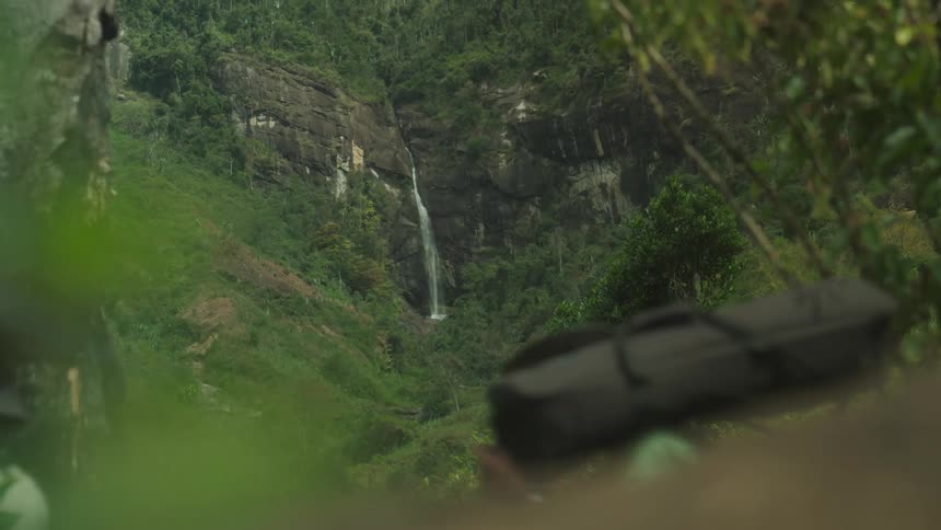 A group of people some carrying supplies walk past a distant waterfall