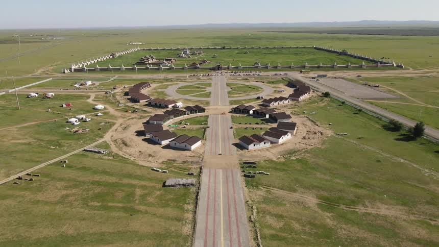 A wide road with a round shaped village and behind it there is a large green lawn surrounded