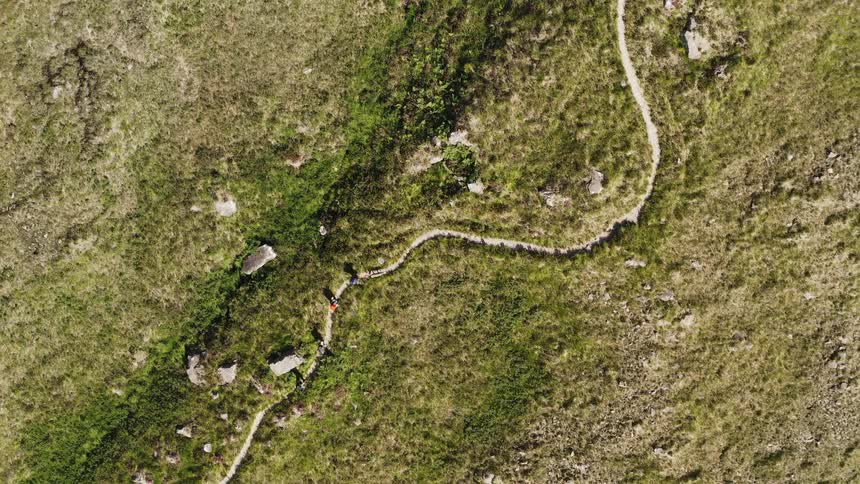 Aerial top view of a small group of hikers walking single file on a trail up a slope surrounded by rocky moss covered terrain