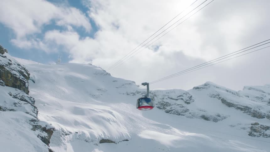 A carriage car glides along a rope high above the ground across the sky above a snowy mountain
