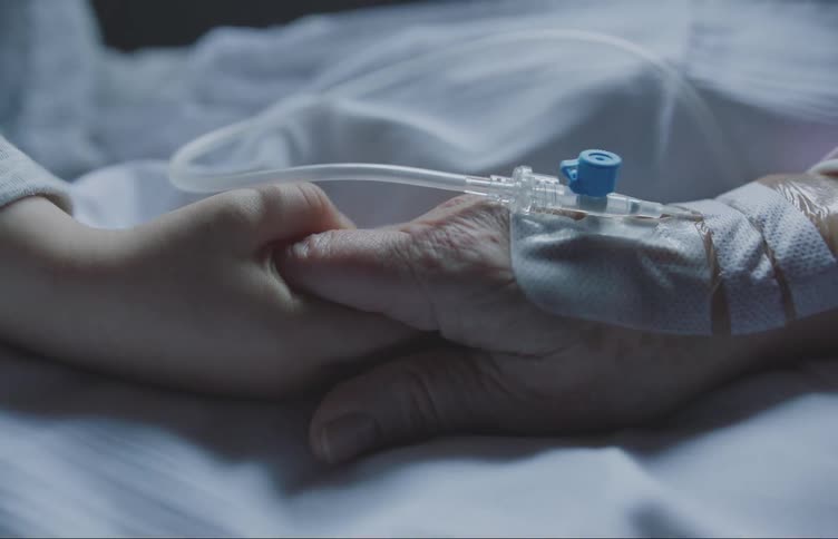 A hand offering comfort to another hand with an iv line set in a dimly lit room