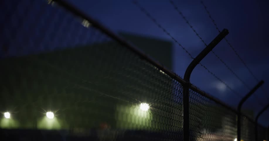 A fence with barbed wire on top around a building with several lights at night