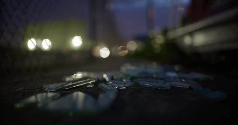 Lights shine through chain link fence onto glass shards lying on the ground at night