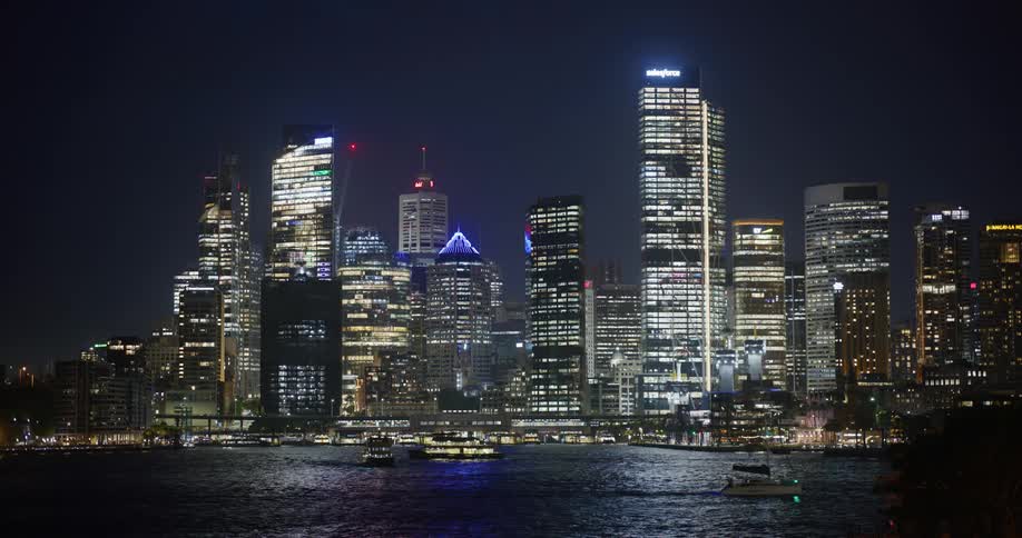 Boats travel through water at night near brightly lit downtown skyscrapers