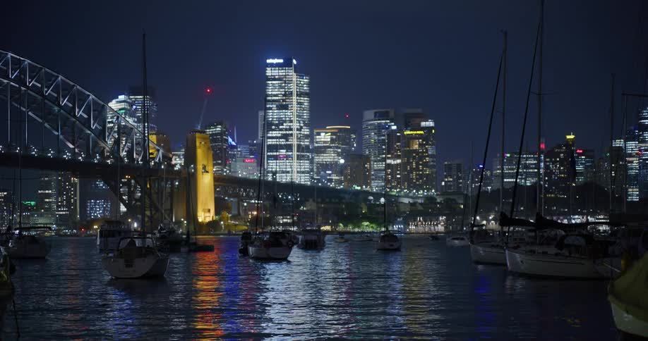 Sailboats float on reflective water across bridge from city skyscrapers at night