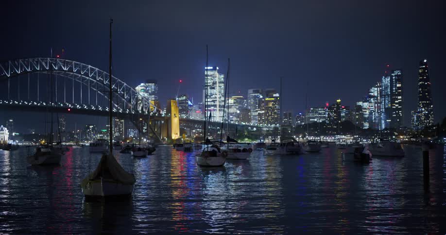View of the sea with boats next to a metropolis with skyscrapers and a bridge at night