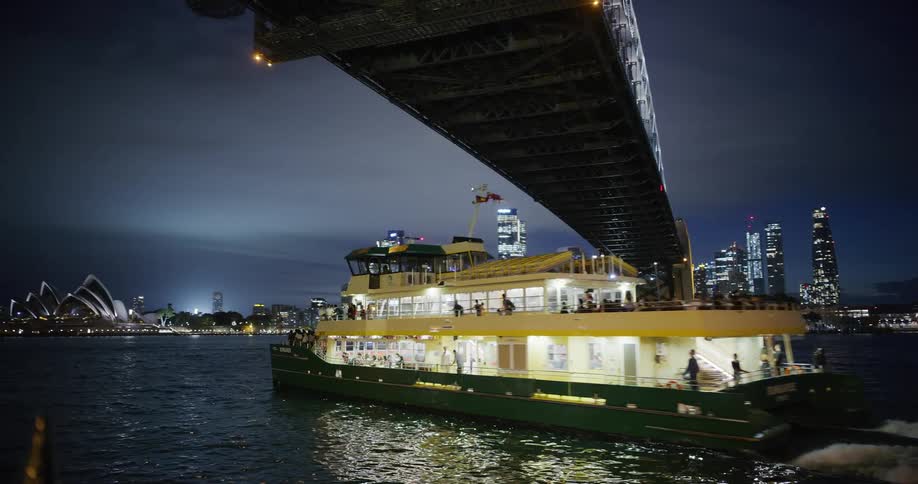 Tourist boat passing under a bridge at night with the sydney skyline in the background