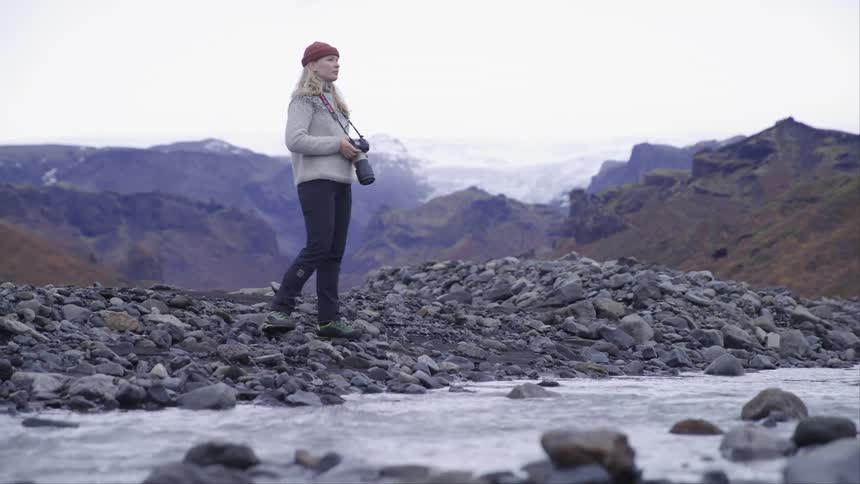 A lady taking pictures with a camera while standing on a rock close to a stream