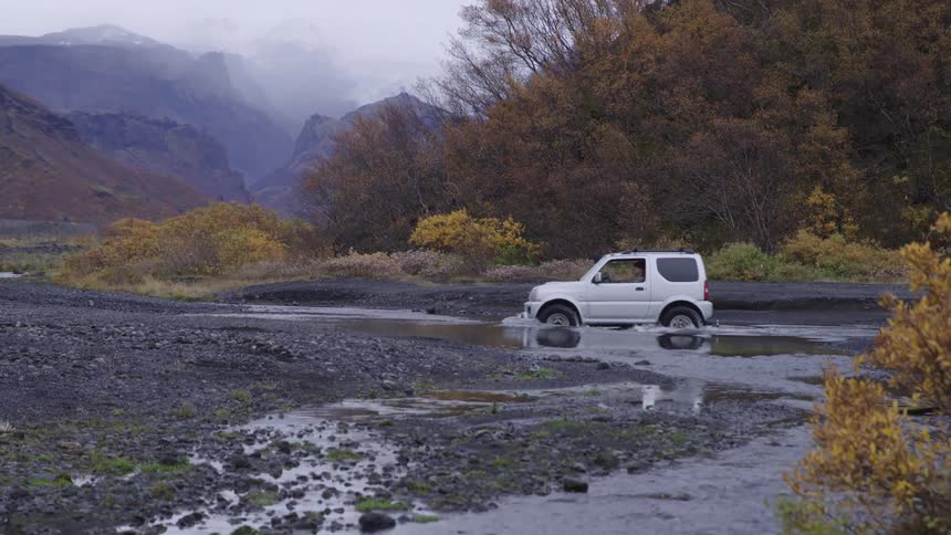 A small suv car driving over some water in a region with forests and mountains