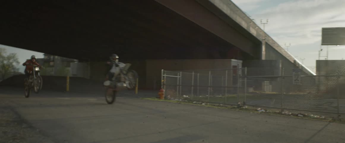 A motorcyclists does a wheelie as he and another motorcyclist pass under a bridge