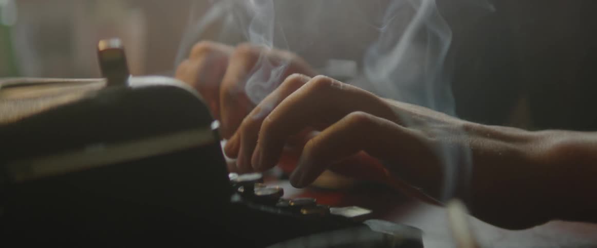 A man typing on a typewriter with a cigarette smoking in the foreground