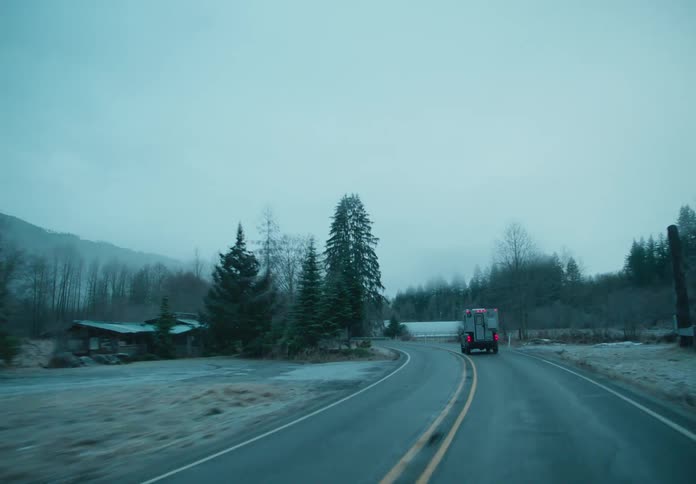 A large car is driving on an asphalt road and there are several trees on the side of the road in a rural region