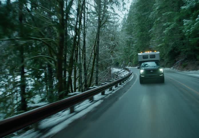 A large car is driving on an asphalt road and there are several trees with snow on the sides of the road