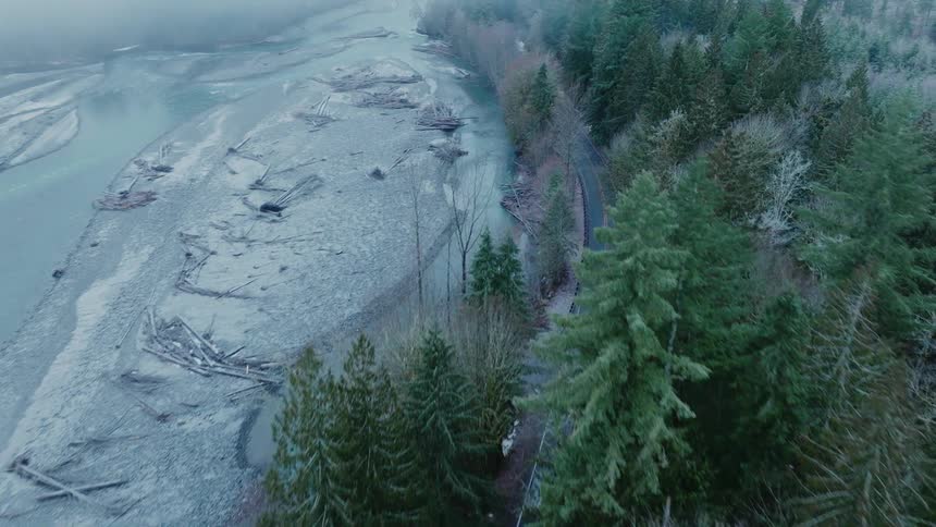 Vehicle follows highway between conifer forest and river during winter