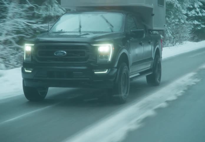 A pickup truck with a camper shell is driving on a road surrounded by dense forest