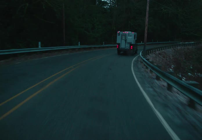 A pickup truck with a camper shell is driving on a road surrounded by dense forest