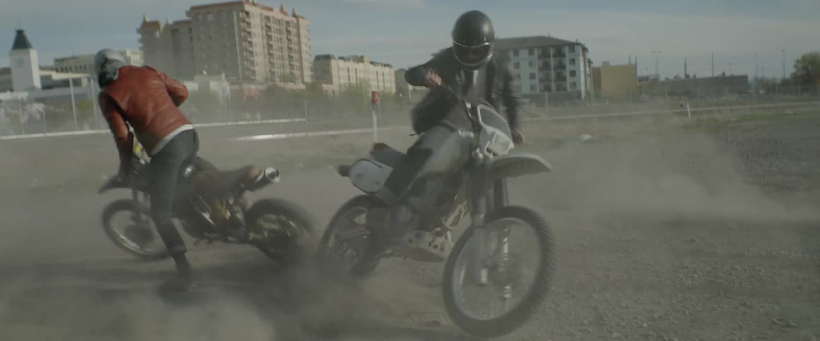 Two people on dirt bikes do tricks in large open dirt area