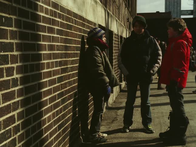 Boys join waiting friend and stand talking near building as others play in the background