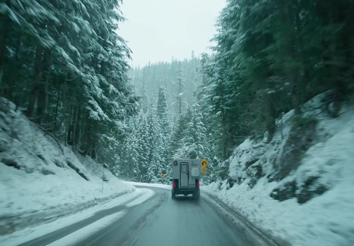Pickup truck with a camper shell drives along highway curving through conifer forest on mountain during winter snowfall
