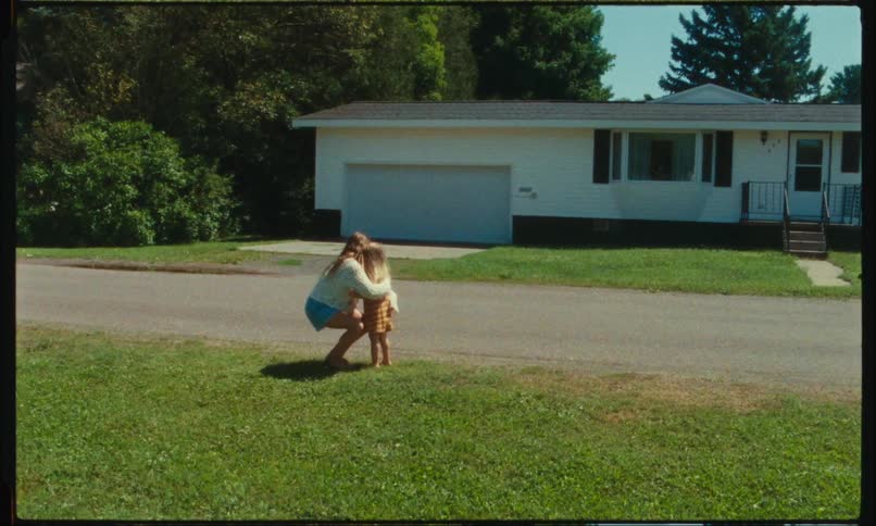 Two girls are standing near a neighborhood street as the big girl bends down and hugs the little girl