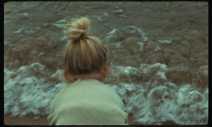 A little girl is standing on the beach and she bends down and puts her hands in the water as waves roll into shore