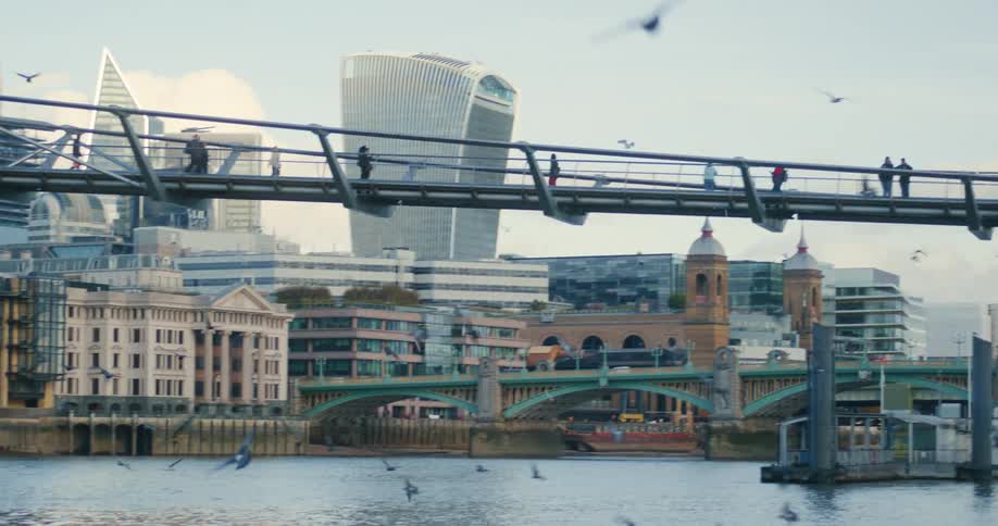 People walk over pedestrian bridge between river bank and downtown buildings as birds fly through the air
