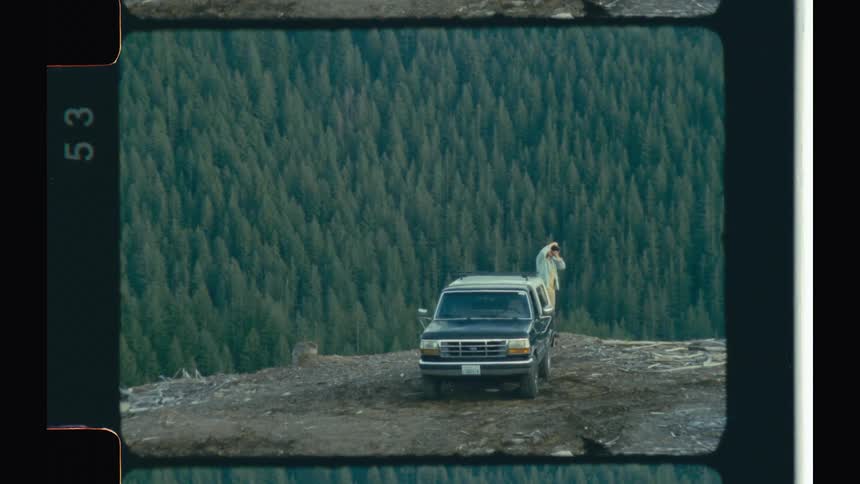 A car and a person parked on a hill and below is a forest of pine trees