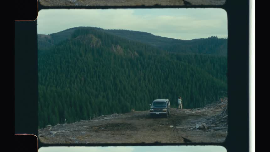 View of a man next to a car on a cliff under a mountain with a scenic backdrop of peaks and forests