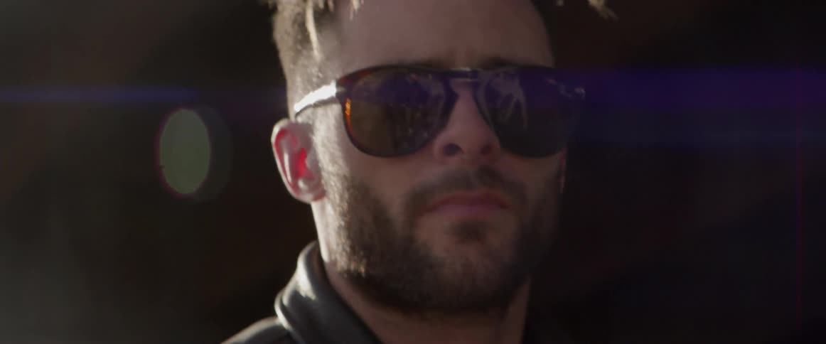 A man wearing sunglasses and a leather jacket turns to look