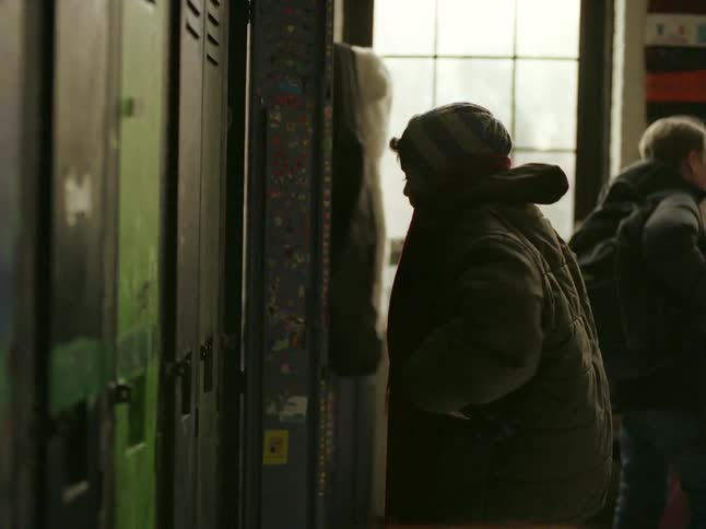 A young child bundled up in warm clothing retrieves items from a locker in a dimly lit hallway while other children in the background converse.